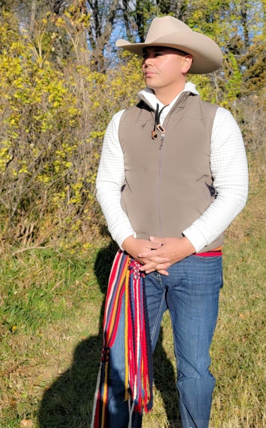 A man stands outdoors, wearing a hat and vest, holding a colorful woven sash.